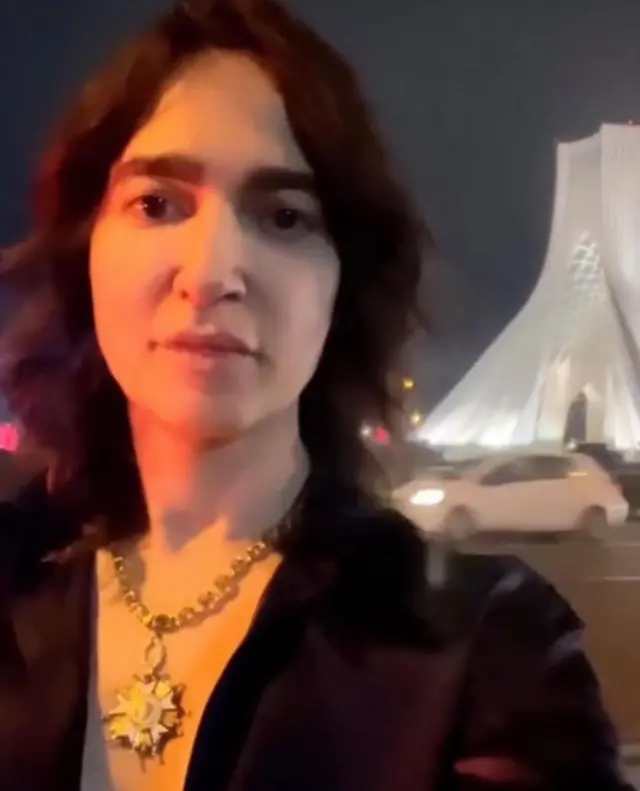 تصویر سرین بدیعی در حالی که پشت سرش برج آزادی و بخشی از میدان آزادی دیده می‌شود. یک ماشین سفید در خیابن دیده می‌شود. سرین بدیعی به دوربین نگاه می‌کند و گردنبند بزرگی به گردن دارد