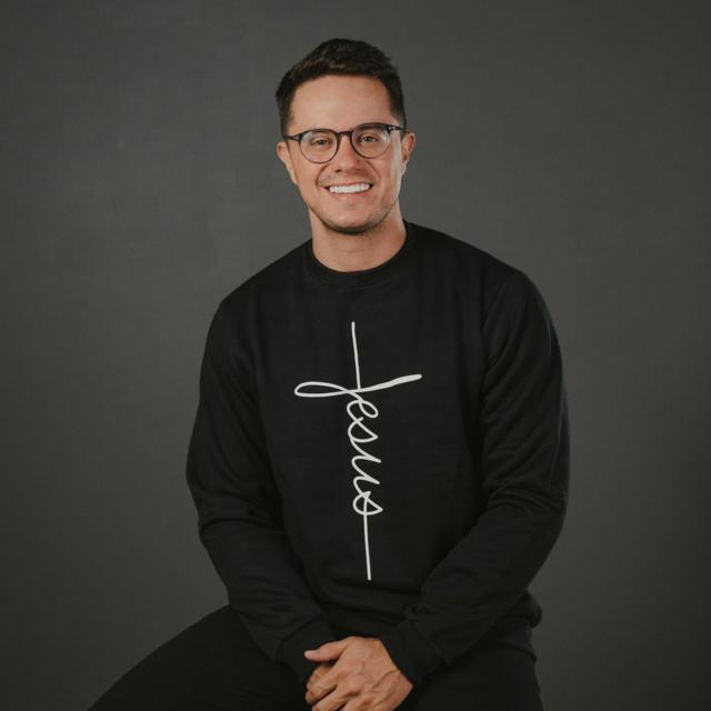 Homem branco de óculos sorrindo com blusa escrito 'jesus'