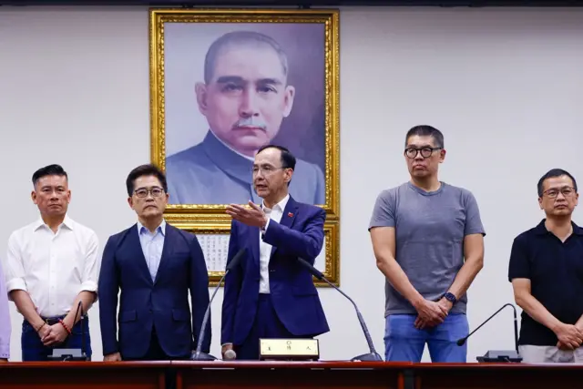 身穿深蓝色西装外套，白色衬衣，左边衣领别上青天白日满地红旗徽章的国民党主席朱立伦（中）在台北中央党部就“大罢免”首轮投票结果向记者发言，国民党领导层成员在其两边站立（26/7/2025）