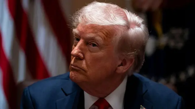 الرئيس الأمريكي دونالد ترامب مرتدياً بدلة رسمية كحلية اللون وخلفه العلم الأمريكي