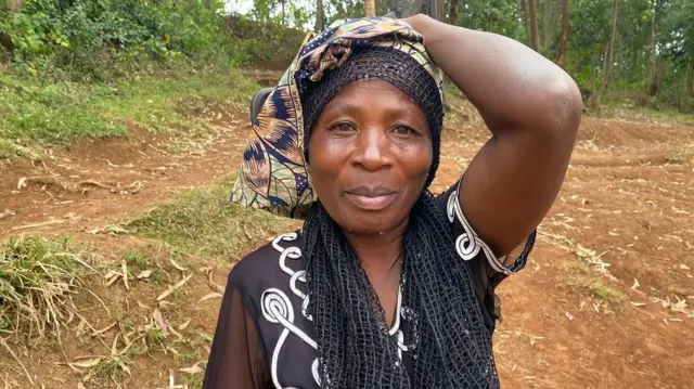 Umugore ushyize ukuboko mu mutwe, afite igitenge n'igitambaro cy'umukara mu mutwe. Yambaye ishati ibonerana y'umukara n'umweru. Arimo kureba muri 'camera'. Inyuma ye hari ibiti n'ibyatsi. 