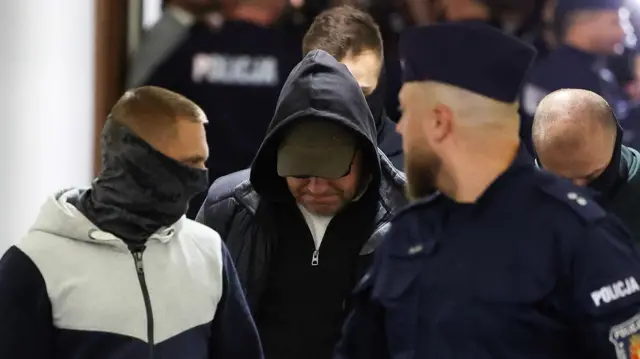 Mężczyzna w czarnej bluzie z kapturem i czapce baseballowej patrzy w dół, a jego oczy są zasłonięte. Eskortują go mężczyzna w mundurze policyjnym i drugi mężczyzna po cywilnemu, który ma na twarzy maskę. W tle widać więcej policjantów.