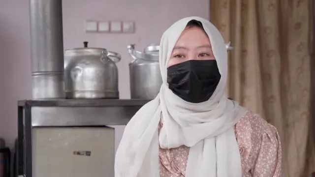 شکیلا با چادر سفید و ماسک سیاه که در عقب‌اش یک بخاری قرار دارد.