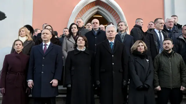 Велика група людей стоїть на східцях перед входом до будівлі теракотового кольору. В першому ряду – зліва направо – Марта та Кароль Навроцькі, Діана та Гітанас Науседа, Олена та Володимир Зеленські. За ними ще кільканадцять людей шикуються щільними рядами для офіційного групового знімка, серед яких видно Андрія Сибігу та Світлану Тихановську.