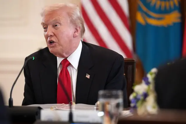 Trump koyu renk takım elbisesiyle Amerikan bayrağı önünde oturmuş konuşma yapıyor