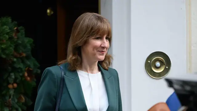 Rachael Reeves tem cabelos na altura dos ombros, um colete verde e uma camisa branca. Ela tem um sorriso no rosto.