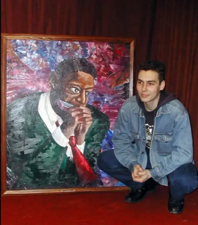 Молодой Андрей Акузин в джинсовке сидит на корточках возле большой картины, на которой темнокожий мужчина играет на губной гармошке