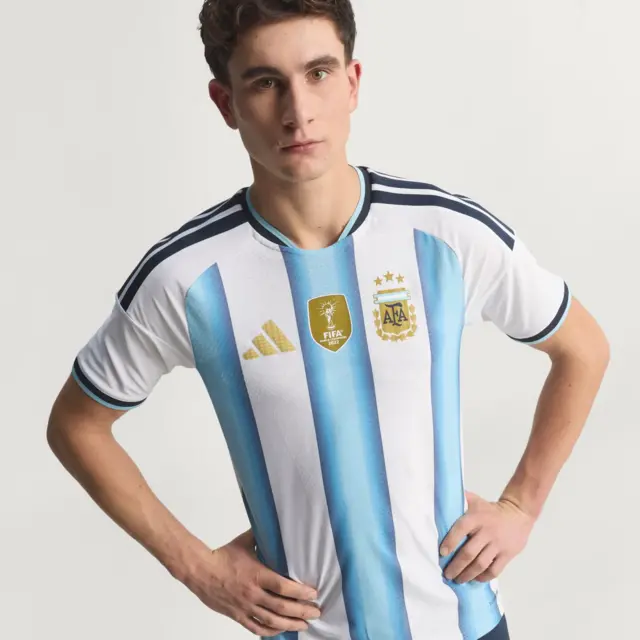 A imagem mostra um jovem usando a camisa listrada em azul-claro e branco da seleção argentina, com detalhes em preto nos ombros e nas mangas. O uniforme exibe o escudo da AFA com três estrelas acima, o selo dourado de campeão mundial da FIFA no centro do peito e o logotipo da Adidas em dourado. O modelo posa com as mãos na cintura e encara a câmera com expressão séria.