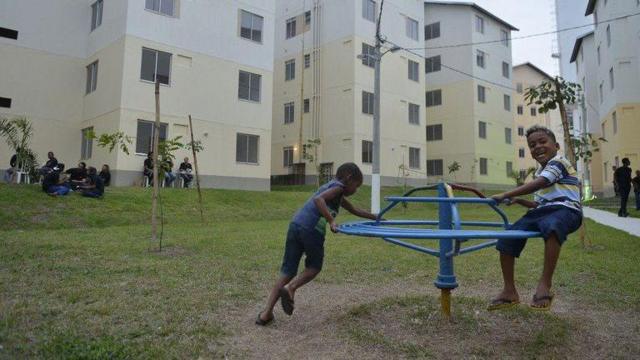 Crianças brincando em gira-gira em frente a edifícios de programa habitacional