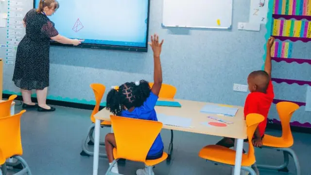 Two children, a girl wey wear blue T-shirt and boy wey wear red T shirt, sit down for orange chairs for one table for classroom. Dem raise dia hands to answer a question wey di teacher wey wear black patterned dress ask, as she dey write on a white board for front of di classroom.