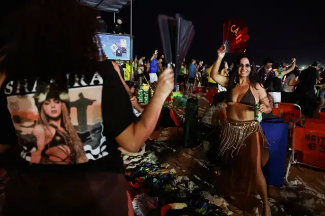 Mulher com leque dançando; outra veste camiseta com rosto e nome de Shakira.