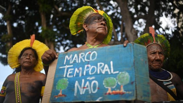 Três homens indígenaswww bet nacional commeia idade durante protesto; um segura uma placa escrito 'marco temporal não'