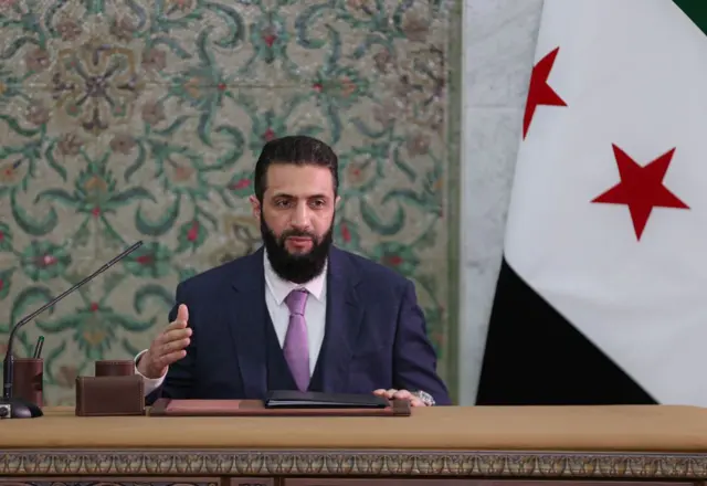الرئيس السوري أحمد الشرع يجلس بجوار علم سوريا خلال إعلان تحديد الدستور المؤقت في دمشق