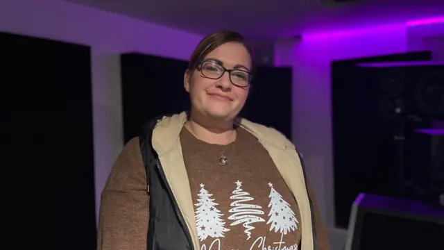 Rebecca Quinn está de pie en la zona de mezclas del estudio. Lleva un jersey navideño marrón con árboles de Navidad blancos y un collar redondo de plata. También lleva un abrigo negro tipo chaleco, desabrochado, que revela un interior beige. Lleva gafas de montura negra y el pelo castaño recogido.