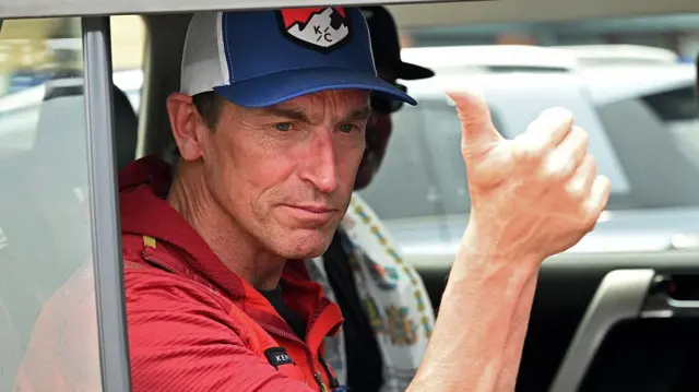 İngiliz dağcı Kenton Cool, kırmızı tişört ve mavi beyzbol şapkası ile iyiyim pozu veriyor, bir araç içinde arka koltukta oturuyor, pencere de açık.