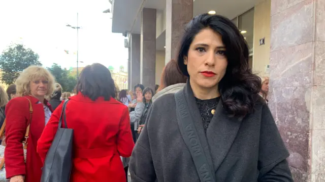 Žena tamne puštene kose sa crvenim karminom u tamnom kaputu gleda u kameru, iza nje je grupa ljudi koja protestuje