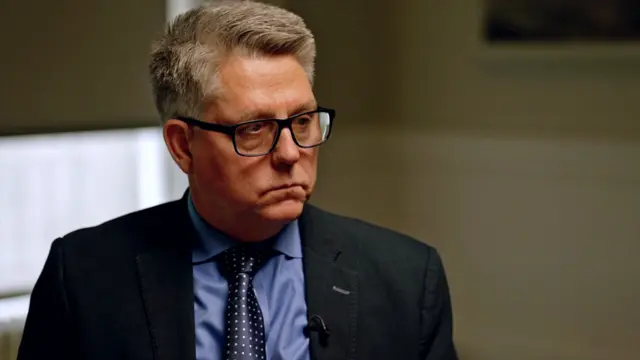 يظهر كيفن هايلاند بشعر أشقر ونظّارات بإطار داكن، مرتدياً بدلة داكنة وقميصاً أزرق وربطة عنق كحلية، في غرفة خافتة الإضاءة وقليلة الزينة، حيث يضيء وجهه نورٌ ناعم بينما يتسرّب الضوء الخارجي عبر ستارة نصف مسدلة.