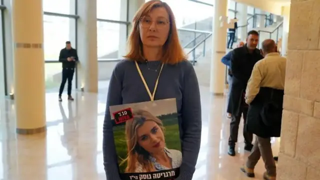 زنی در سالن بزرگ ساختمانی ایستاده و تصویر دختر کشته شده‌اش به دست حماس را به گردن آویخته