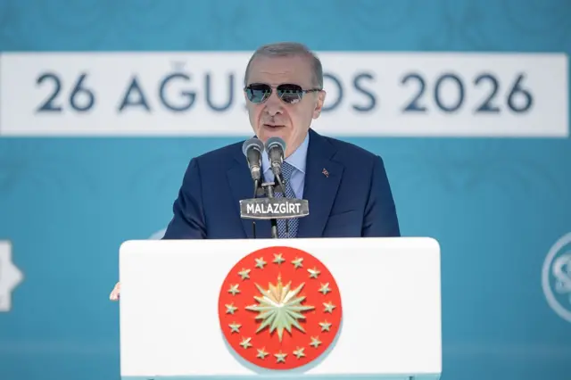 Erdoğan turkuaz arka planda bir kürsüde güneş gözlükleriyle konuşma yapıyor.