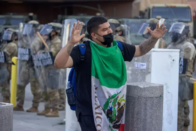 Un hombre envuelto en una bandera de México con los brazos en alto camina frente a soldados de la Guardia Nacional de California.