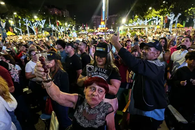 Una mujer con una diadema que dice "Feliz Año Nuevo" alza una copa entre una multitud que celebra la llegada de 2026 en las calles de Caracas