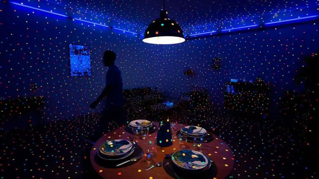 Sala escura com pontos de luz coloridos projetados por toda parte, incluindo nas paredes, no teto, no chão e sobre uma mesa redonda com pratos, taças e utensílios para quatro pessoas.