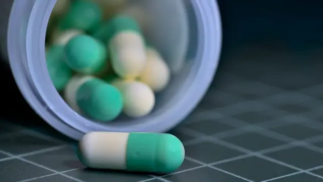 حبوب أدوية بلونين أبيض وأخضر داخل علبة مفتوحة، وهناك حبة واحدة خارج العلبة البلاستيكية الشفافة. على أرضية تميل إل اللون الرمادي ومخطط بالأبيض.
