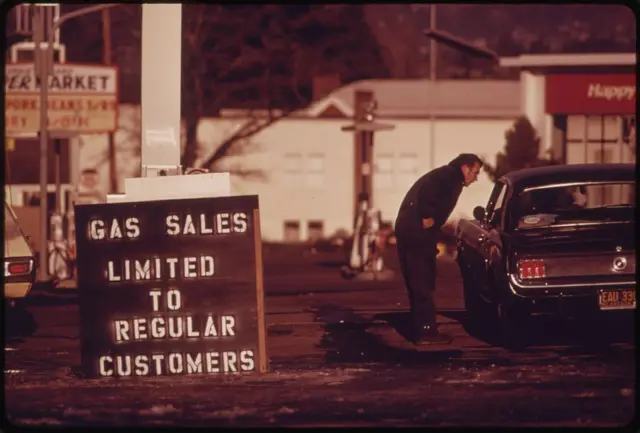 An image of a billboard outside a petrol station in the US during the 1973 oil crisis, saying 'Gas Sales Limited to Regular Customers'. In the background a man can be seen leaning in to a car window and talking to the driver.
