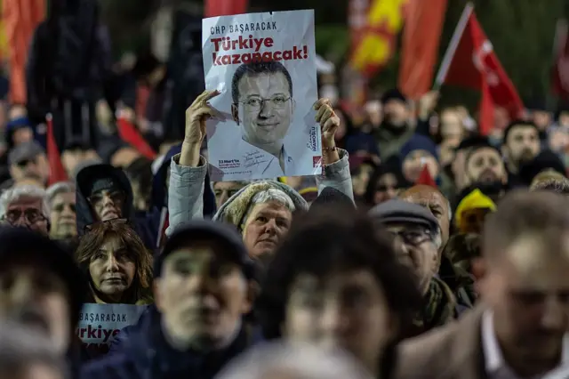18 Mart'ın ylı dönümünde toplanan kalabalık, bir kadının elinde İmamoğlu'nun fotoğrafının bulunduğu ve "Türkiye kazanacak" yazan bir pankart var. 