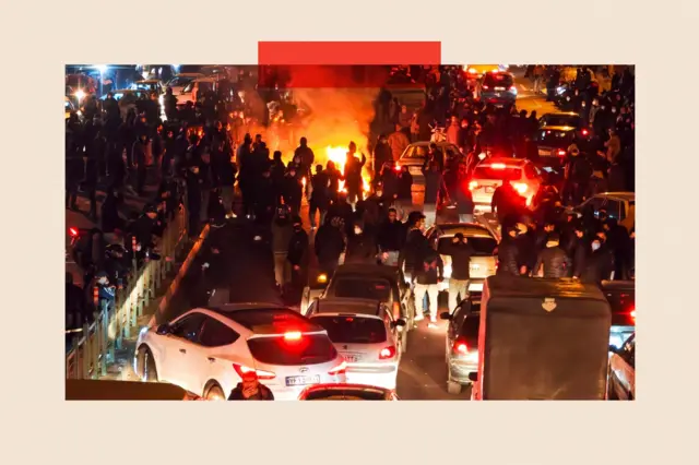 Sokaklarda protesto gösterisi yapan yüzlerce insan, uzakta bir ateş yakılmış halde ve ön planda arabalar görülüyor.