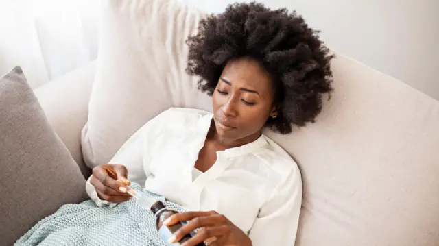 Woman dey drink cough syrup ontop bed inside house.