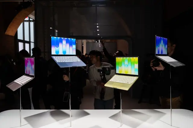 Четыре ноутбука MacBook Neo с заставками разных цветов стоят на столбиках, прикрепленных к светлой горизонтальной поверхности. Их фотографируют люди.