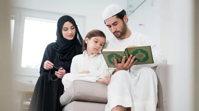 أبوان يجلسان بجوار ابنهما الصغير، إذ يقرأ الأب القرآن في رمضان.