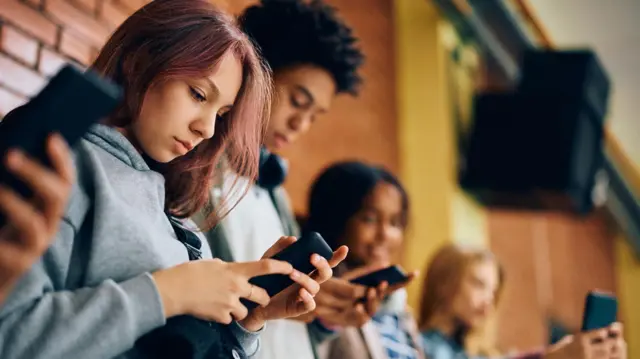 Quatro alunos de pé contra uma parede de tijolos, sem falar uns com os outros enquanto cada um olha para um telefone em suas mãos