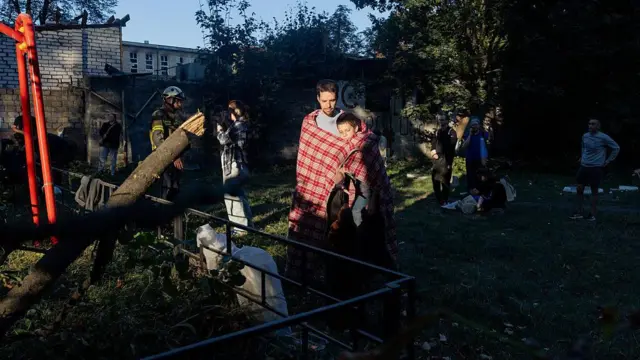 Мужчина, закутавшись в плед в бело-красную клетку, держит на руках мальчика лет шести, у его ног стоит собака среднего размера.