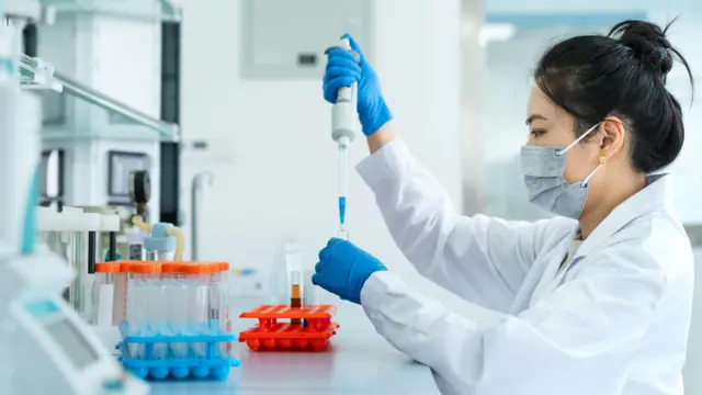 Un entorno de laboratorio donde una persona con bata blanca trabaja en una mesa de laboratorio. La persona lleva guantes protectores y mascarilla mientras utiliza cuidadosamente una pipeta para transferir líquido a un pequeño tubo de ensayo. En primer plano, sobre la superficie de trabajo, hay varias gradillas con tubos de ensayo que contienen líquidos de colores, dispuestas de forma ordenada. Al fondo se aprecian equipos científicos y estanterías, lo que sugiere un espacio de trabajo limpio y organizado.