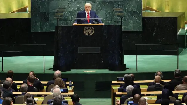 Tổng thống Mỹ Donald Trump, mặc bộ vest màu xanh đậm, áo sơ mi trắng và cà vạt đỏ, phát biểu trước Đại hội đồng Liên Hợp Quốc, với các thành viên ngồi tại bàn, từ bục phát biểu có biểu tượng của Liên Hợp Quốc, trên một sân khấu màu xanh lá cây, tại New York 