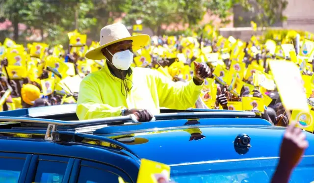 Prezida Yoweri Museveni yambaye inkofero , agapfumunwa n'ishati y'umuhondo; akaba ahagaze mu modoka