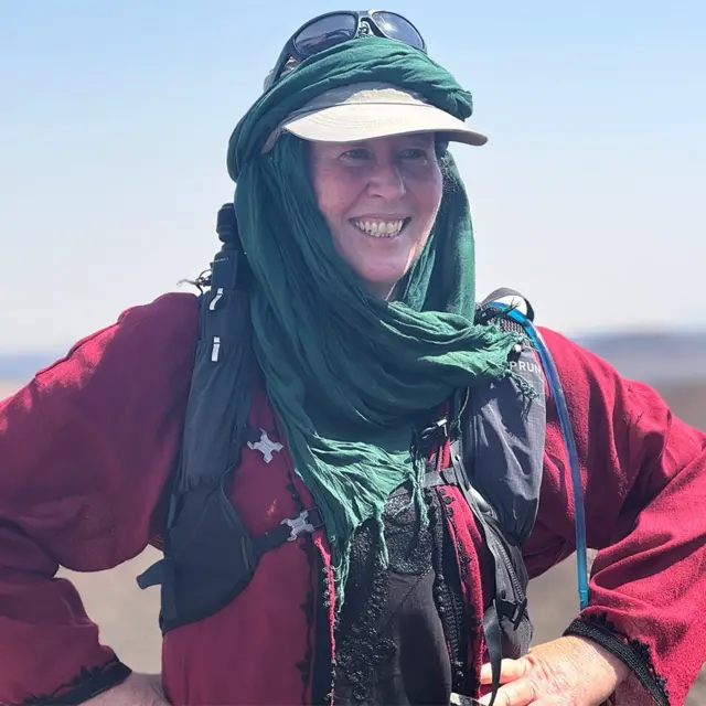 Alice Morrison, con un pañuelo en la cabeza, una gorra y una mochila, de pie al aire libre en un paisaje desértico 