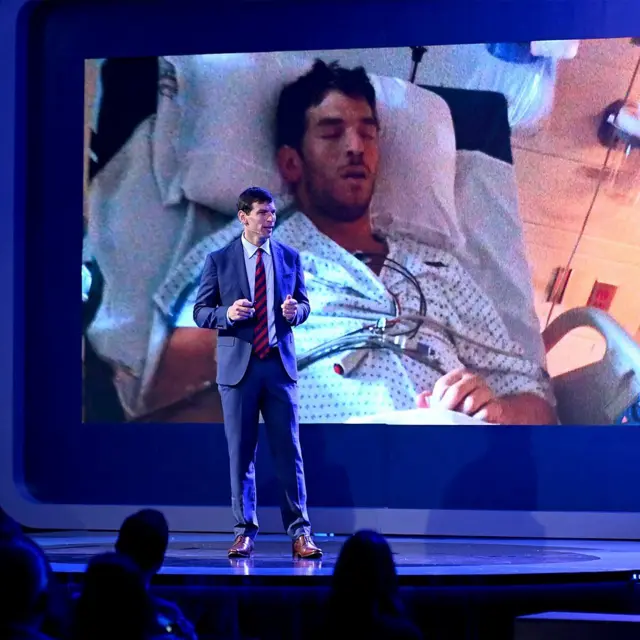 David Fajgenbaum em uma palestra. Ao fundo, imagem de um homem no hospital.