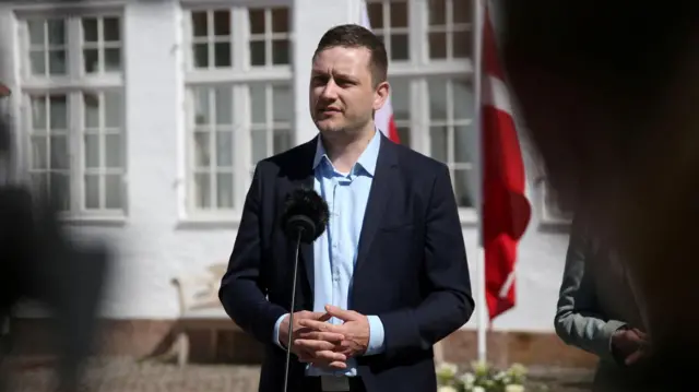 Jens-Frederik Nielsen, Greenland’s Prime Minister, wears a navy blue suit and a light blue shirt as he speaks to the press from behind a microphone. The Greenlandic and Danish flags are visible behind him.