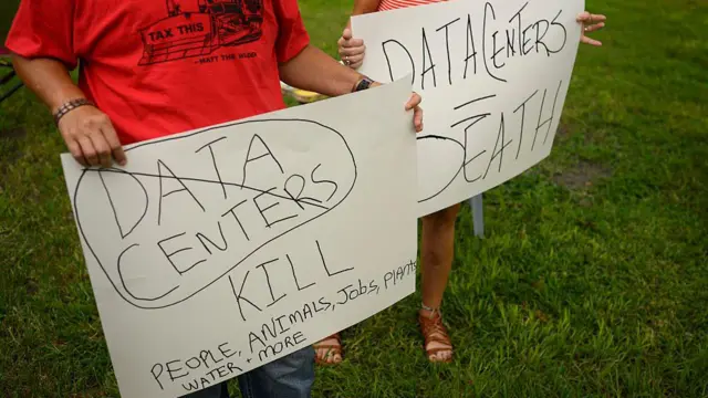 شخصان يرفع كل منهما لافتة كتب على إحداها Data centers=death (مراكز البيانات=الموت)، وعلى الأخرى data centers kill people, animals, jobs, plants, water and more (مراكز البيانات تقضي على البشر والحيوانات والوظائف والنباتات والمياه وعيرها)