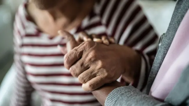 Duas pessoas de mãos dadas onde apenas as mãos aparecem; possivelmente um paciente idoso e um cuidador que o auxilia.
