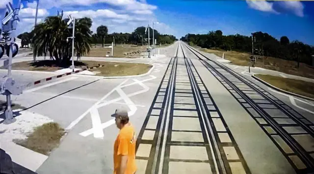 Randy Johanson, con camiseta naranja y gorra, captado por las cámaras del tren instantes antes de que lo arrollara, Las vallas del paso a nivel están bajadas. 