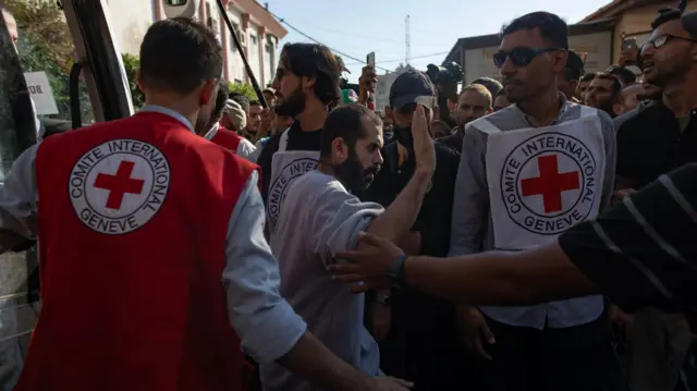 Un hombre con barba saluda rodeado de representantes de la Cruz Roja Internacional, que visten chalecos con el logo de la organización.