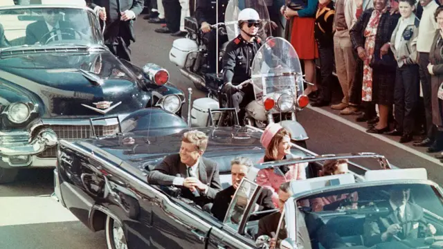 1963 年 11 月 22 日，美国总统约翰·F·甘迺迪、第一夫人杰奎琳·甘迺迪、德州州长约翰·康纳利（John Connally）等人在德州达拉斯向车队沿途的人群微笑。几分钟后，总统的汽车经过迪利广场时遇刺身亡。