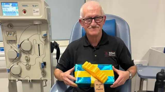 Un hombre sentado en una silla en un consultorio médico, con una máquina de donación de sangre a su lado. Lleva gafas y camisa negra. Sonríe mientras sostiene un regalo azul envuelto en un lazo amarillo con una etiqueta marrón que dice "mi regalo de plasma".