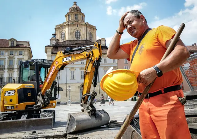 Un trabajador de la construcción, vestido con un uniforme naranja de alta visibilidad, sostiene un casco amarillo y se seca la cabeza frente a una excavadora, junto a una plaza en Turín (Italia) el día 25 de junio.
