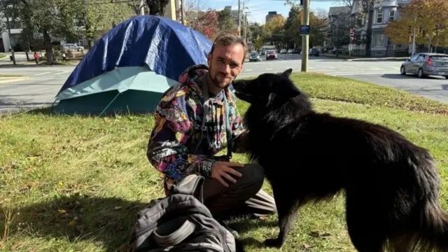 Homem agachado ao ladoesporte uolcachorro,esporte uolgramado com barraca ao fundo,esporte uolzona urbana