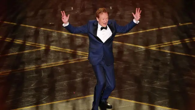 Конан О' Брајан на сцени у тегет оделу, белој кошуљи и црном лептир машном